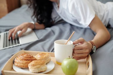 Yatakta yatıp dizüstü bilgisayar kullanan, evde çalışırken yatakta kahvaltı yapan tanınmayan bir adamın yakın çekimi.