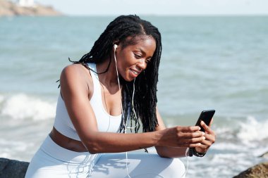 Gülümseyen bayan koşucu plajda antrenman yaptıktan sonra dinleniyor ve aile üyelerine video çağrısı yapıyor.