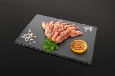 Izgara karides. Siyah bir tahta üzerinde. Bitkilerle ve limon dilimleriyle süslenmiş. Karanlık arka plan. İzole edilmiş. Yakın plan..