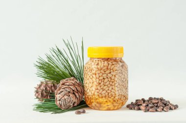 Sarı kapaklı şeffaf bir kavanozda ballı çam fıstığı. Yakınlarda çam kozalakları, çam iğneleri ve fındık var. Etiket için bir yer. Yakın plan. Hafif arkaplan.