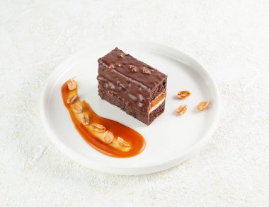 Snickers cake. Dessert based on chocolate biscuit, creamy mousse with caramel. Decorated with caramelized peanuts. On a white plate. Light background. View from above