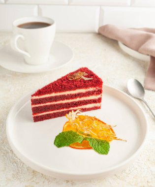 Kırmızı kadife kek. Beyaz tabakta bir parça. Karamel ve kurutulmuş portakalla süslenmiş. Arka planda, bir fincan kahve. Hafif arkaplan.