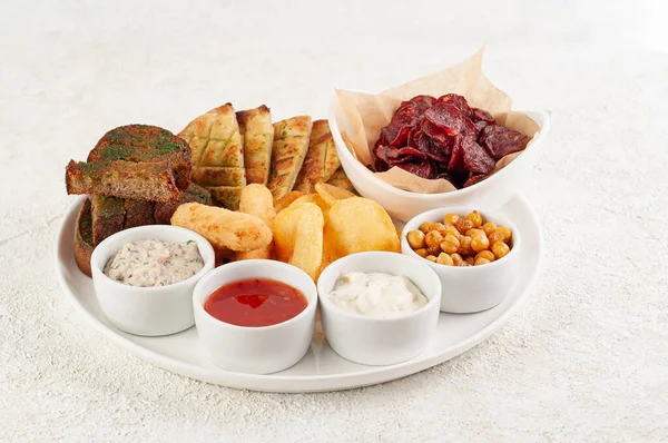 A set of snacks for beer. From croutons, pita bread, smoked sausage and nuggets. Served with three types of sauces. On a white plate. Light background.