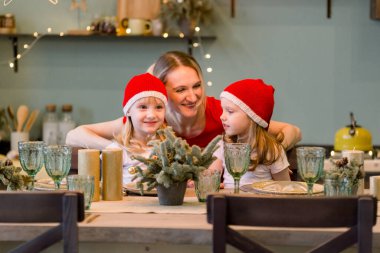 Noel Baba şapkalı, neşeli iki küçük kızla festival masasında oturan annem. Noel zamanı. Yeni yıl 2021. Kızlarıyla eğlenen bir kadın.