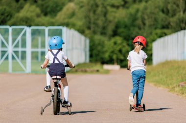 Bisiklet ve scooterla eğlenen iki küçük kızın arka planı. Miğferli neşeli kız kardeşler dışarıda geziyorlar..