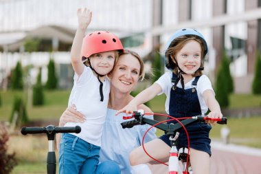 İki güzel kızı olan anne eğleniyor. Bisiklet ve scooter süren kasklı kadın ve kızlar.
