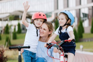 İki güzel kızı olan anne eğleniyor. Bisiklet ve scooter süren kasklı kadın ve kızlar.