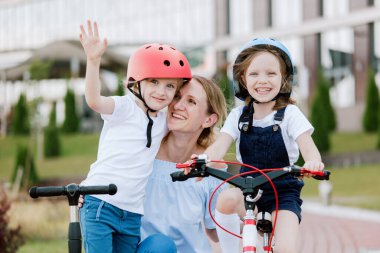 İki güzel kızı olan anne eğleniyor. Bisiklet ve scooter süren kasklı kadın ve kızlar.