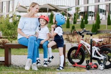 İki güzel kızı olan anne eğleniyor. Bisiklet ve scooter süren kasklı kadın ve kızlar.