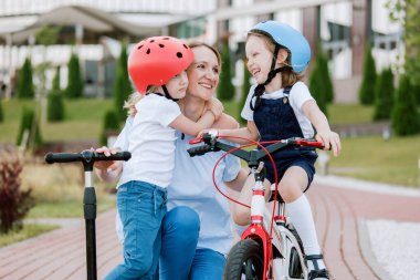 İki güzel kızı olan anne eğleniyor. Bisiklet ve scooter süren kasklı kadın ve kızlar.