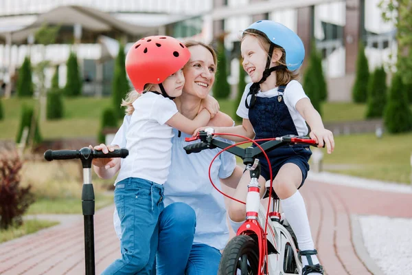 İki güzel kızı olan anne eğleniyor. Bisiklet ve scooter süren kasklı kadın ve kızlar.
