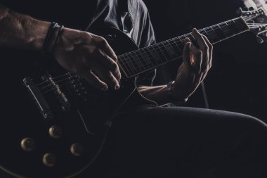 Tişörtlü adam arka planda elektro gitar çalıyor.