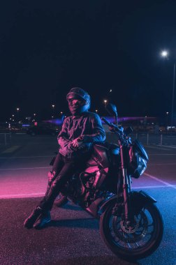 Motosikletçi, gece vakti boş bir otoparkta, neon ışıkta motosikletin üzerinde oturur.