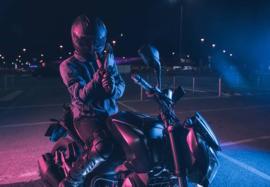Motosikletçi, gece vakti boş bir otoparkta, neon ışıkta motosikletin üzerinde oturur.