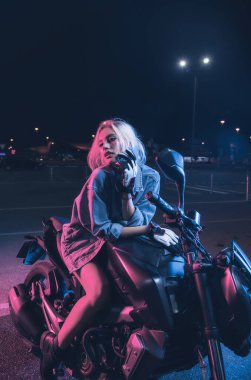 Boş bir otoparkta, gece vakti bir motosikletin üzerinde neon ışıklı bir kızın portresi.