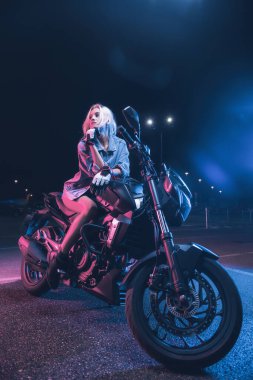 Boş bir otoparkta, gece vakti bir motosikletin üzerinde neon ışıklı bir kızın portresi.