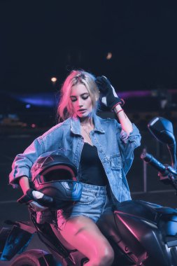 Boş bir otoparkta, gece vakti bir motosikletin üzerinde neon ışıklı bir kızın portresi.
