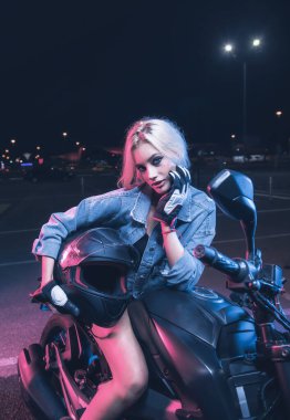 Boş bir otoparkta, gece vakti bir motosikletin üzerinde neon ışıklı bir kızın portresi.