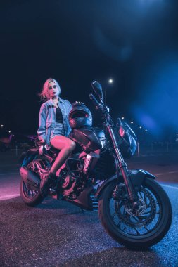 Boş bir otoparkta, gece vakti bir motosikletin üzerinde neon ışıklı bir kızın portresi.