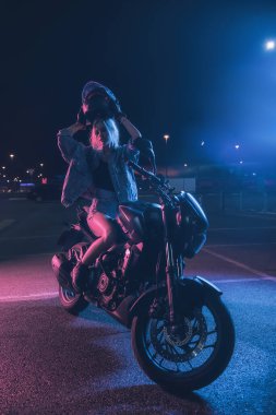 Boş bir otoparkta, gece vakti bir motosikletin üzerinde neon ışıklı bir kızın portresi.