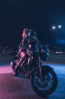 Boş bir otoparkta, gece vakti bir motosikletin üzerinde neon ışıklı bir kızın portresi.