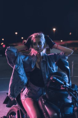 Boş bir otoparkta, gece vakti bir motosikletin üzerinde neon ışıklı bir kızın portresi.