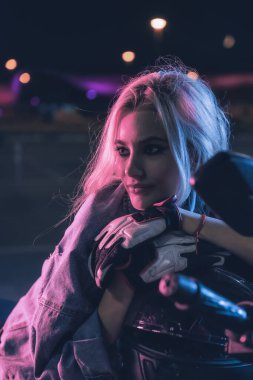 Boş bir otoparkta, gece vakti bir motosikletin üzerinde neon ışıklı bir kızın portresi.