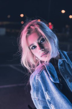 Boş bir otoparkta, gece vakti bir motosikletin üzerinde neon ışıklı bir kızın portresi.