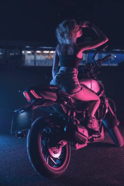 Boş bir otoparkta, gece vakti bir motosikletin üzerinde neon ışıklı bir kızın portresi.