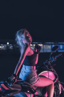 Boş bir otoparkta, gece vakti bir motosikletin üzerinde neon ışıklı bir kızın portresi.