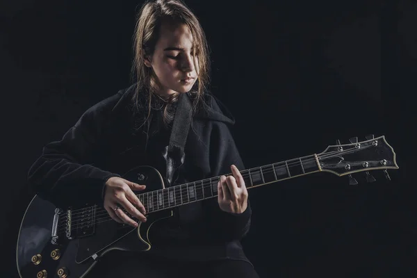 穿着黑色运动衫的女孩在深色背景下弹奏电吉他 图库图片