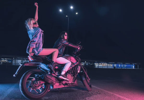 a couple of girls in love on a motorbike in the rays of neon light in an empty parking lot at night