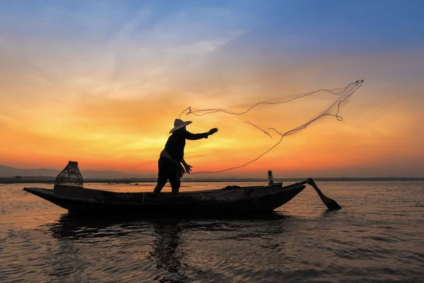 Silueta de pescador asiático en barco de madera en acción lanzando una red para la captura de peces de agua dulce en el río naturaleza en la madrugada antes del amanecer —  Fotos de Stock