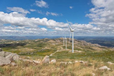 Rüzgâr türbinleri, yeşil ot tarlaları ve kirli yollarla kaplı bulutlu bir gökyüzü manzarası