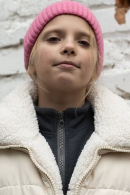 Pembe bereli ve yün yelekli bir çocuğun düşük açılı portresi kendinden emin bir bakışla aşağıya bakıyor, cesur bir gençlik tavrı yakalamış.