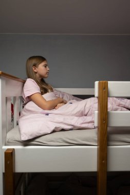 Genç kız ranzada oturuyor, elleri battaniyenin üzerinde dinleniyor, yan yana bakıyor, yumuşak akşam ışığı sakin, sessiz pastel renkler yaratıyor.