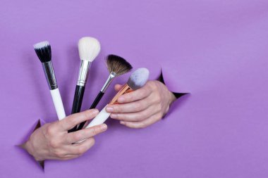 Ellerinizde profesyonel makyaj fırçaları, mor bir arka planda delikler. Metin için boşluk.