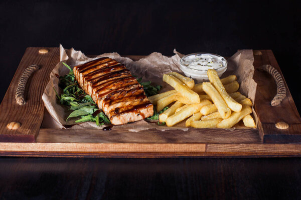 salmon fillet steak with french fries on a wooden tray, beautiful serving, dark background.