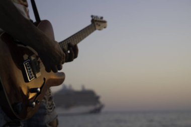 Günbatımında elektro gitar çalan müzisyen, arka planda yolcu gemisi