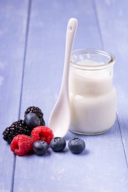 in the foreground, on the wooden table, some fresh berries and a jar of plain and low-fat yogurt with a ceramic spoon