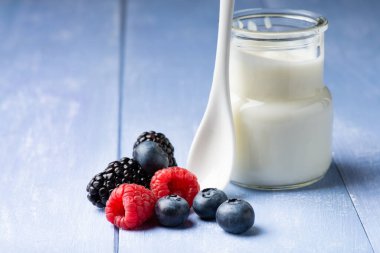 in the foreground, on the wooden table, some fresh berries and a jar of plain and low-fat yogurt with a ceramic spoon