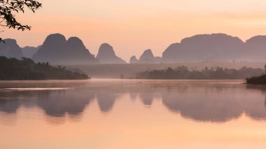 Gün doğumunda sakin bir yaz gölü. Dramatik dalga bulutları ve gündoğumu göle yansıyor. Arka planda dağlar ve tropikal ormanlar var. Nong Talay, Tayland. Göle hafifçe odaklan..