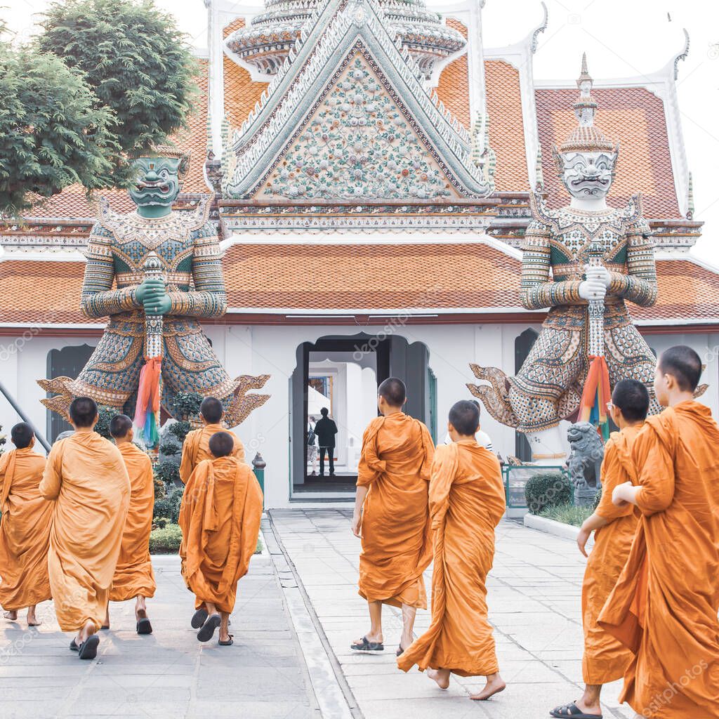 A group of Buddhist monks and novices visiting Wat Arun (Temple of Dawn), the sculptures of two mythical giant demons blurred in the background. Famous place in Bangkok, Thailand.