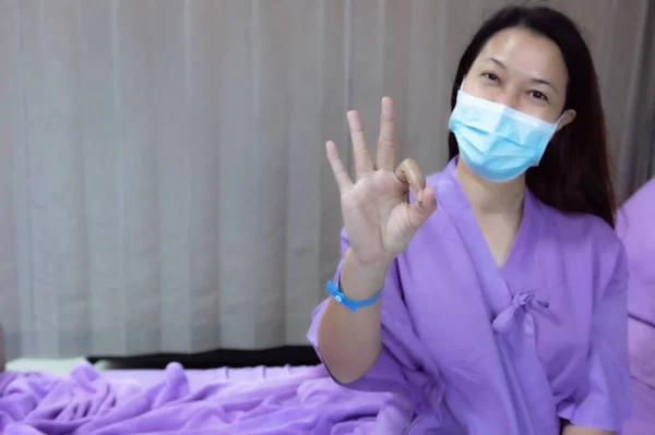 Young Asian female patient is smiling and showing ok gesture. Patient feels happy and comfortable with treatment and therapy on hospital bed in hospital room. Medical healthcare concept.