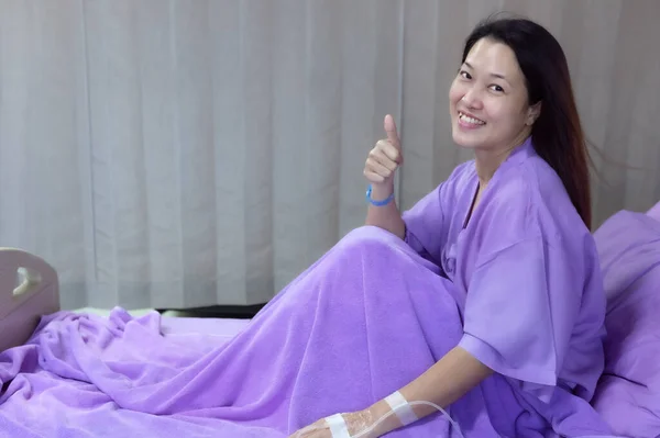 Young Asian female patient is smiling and showing Thumbs up gesture. Patient feels happy and comfortable with treatment and therapy on hospital bed in hospital room. Medical healthcare concept.