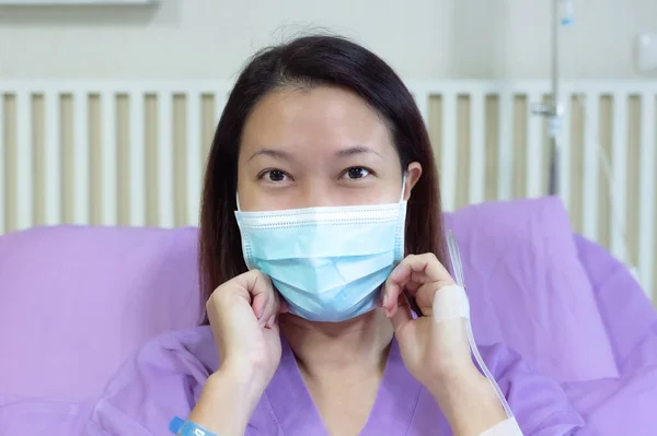 Young Asian female patient is smiling and showing medical mask. Patient feels happy and comfortable with treatment and therapy on hospital bed in hospital room. Medical healthcare concept.
