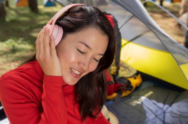 Kırmızı elbiseli güzel bir kadın kulaklıklardan müzik dinlemenin keyfini çıkarıyor. O, sarı bir çadırda oturdu. Pang Oung, Mae Hong Son, Tayland Gölü 'nün yanındaki güzel bir çam ormanının ortasında kamp yapan güzel doğa manzarasına hayranım.