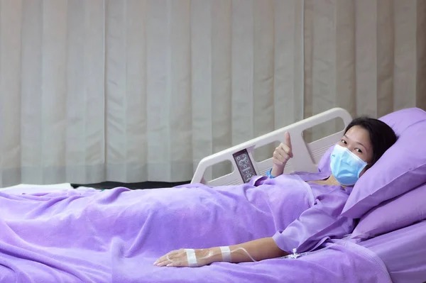 Young Asian female patient is smiling and showing Thumbs up gesture. Patient feels happy and comfortable with treatment and therapy on hospital bed in hospital room. Medical healthcare concept.