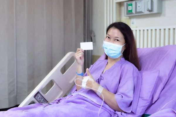 Young Asian female patient is smiling and showing blank credit card, Patient feels happy and comfortable with treatment and therapy on hospital bed in hospital room. Healthcare and Insurance concept.