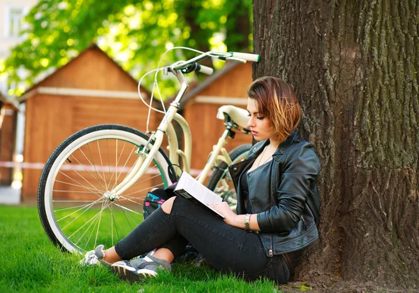 Mladá žena s knihou čtení jízdních kol v městském parku na trávě — Stock fotografie